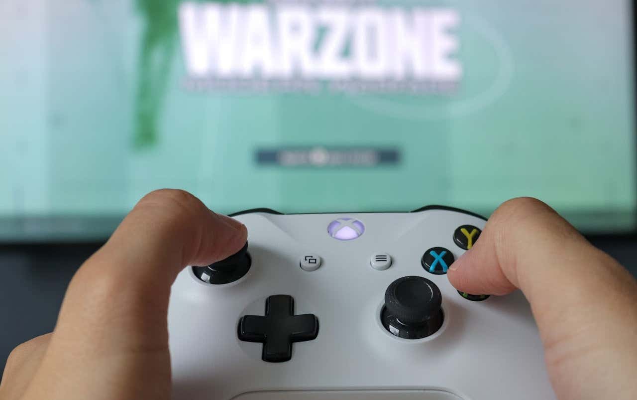 A person holding a white Xbox controller with the game title "WARZONE" displayed on a blurred TV screen in the background